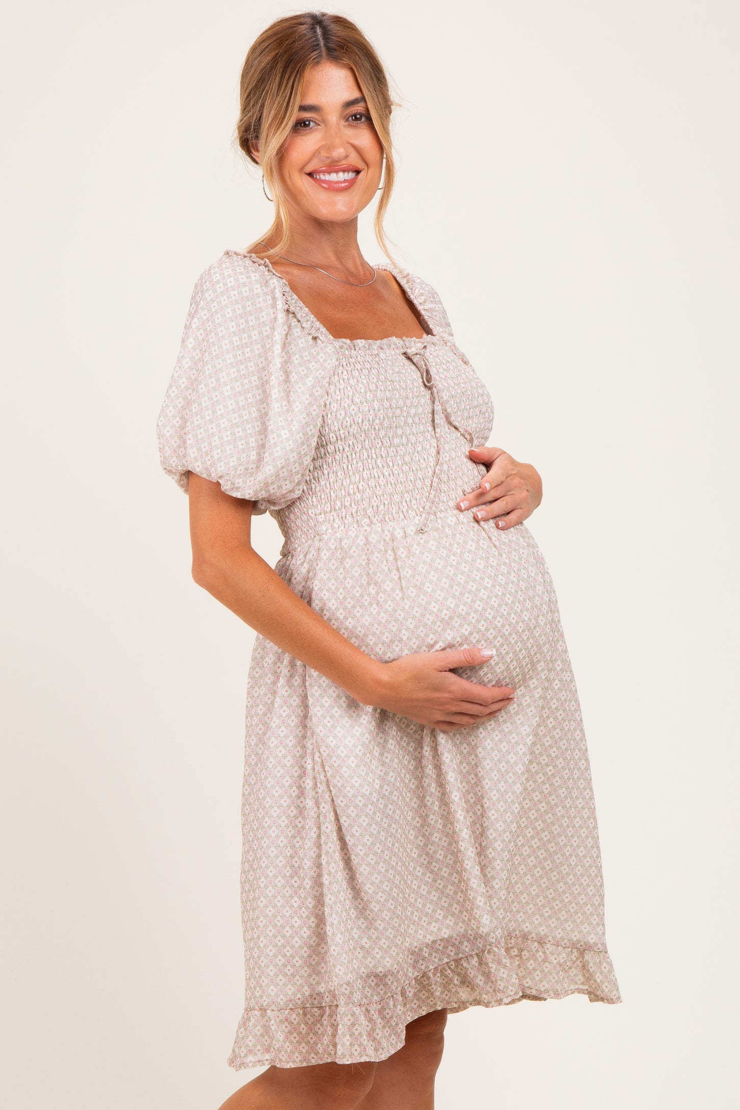 woman wearing a light beige maternity dress with smocked bodice and puff sleeves, gently cradling her baby bump