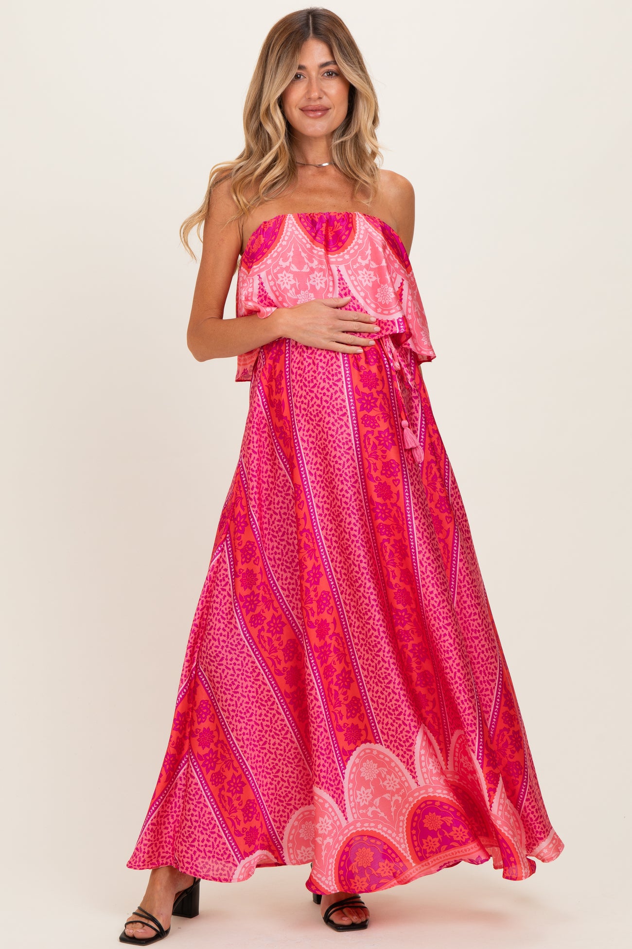 woman wearing a vibrant pink strapless maternity dress with a ruffled overlay and holding her baby bump