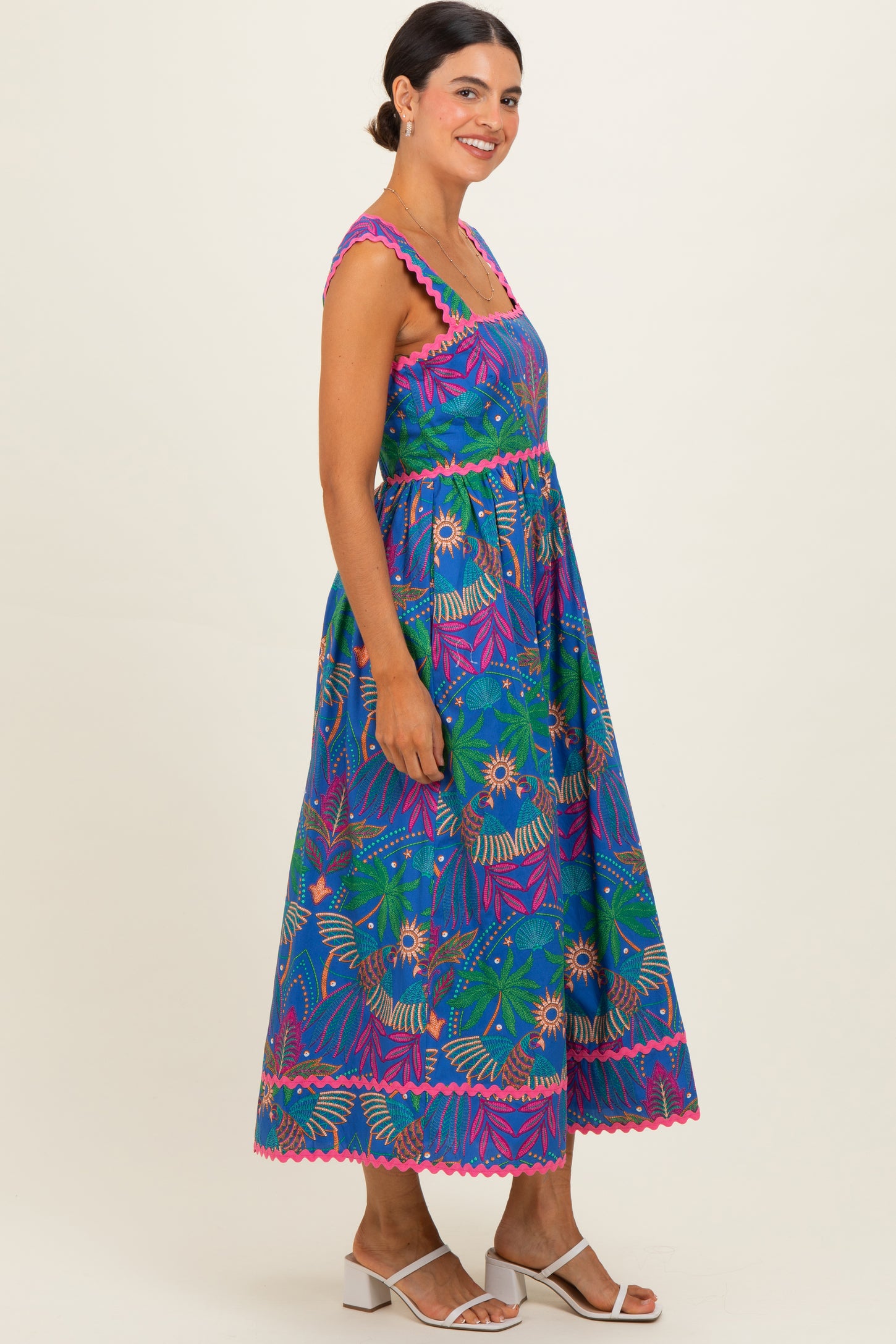 woman wearing a colorful sleeveless maxi dress with floral patterns and scalloped details, standing sideways