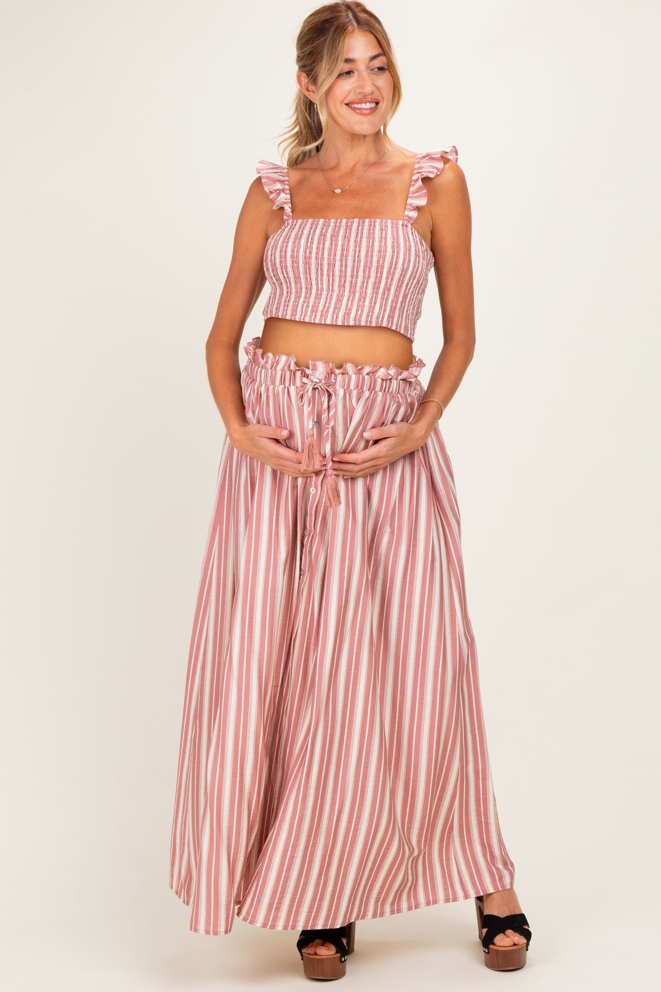 woman wearing a pink and white striped maternity two-piece set with ruffled straps and holding her baby bump