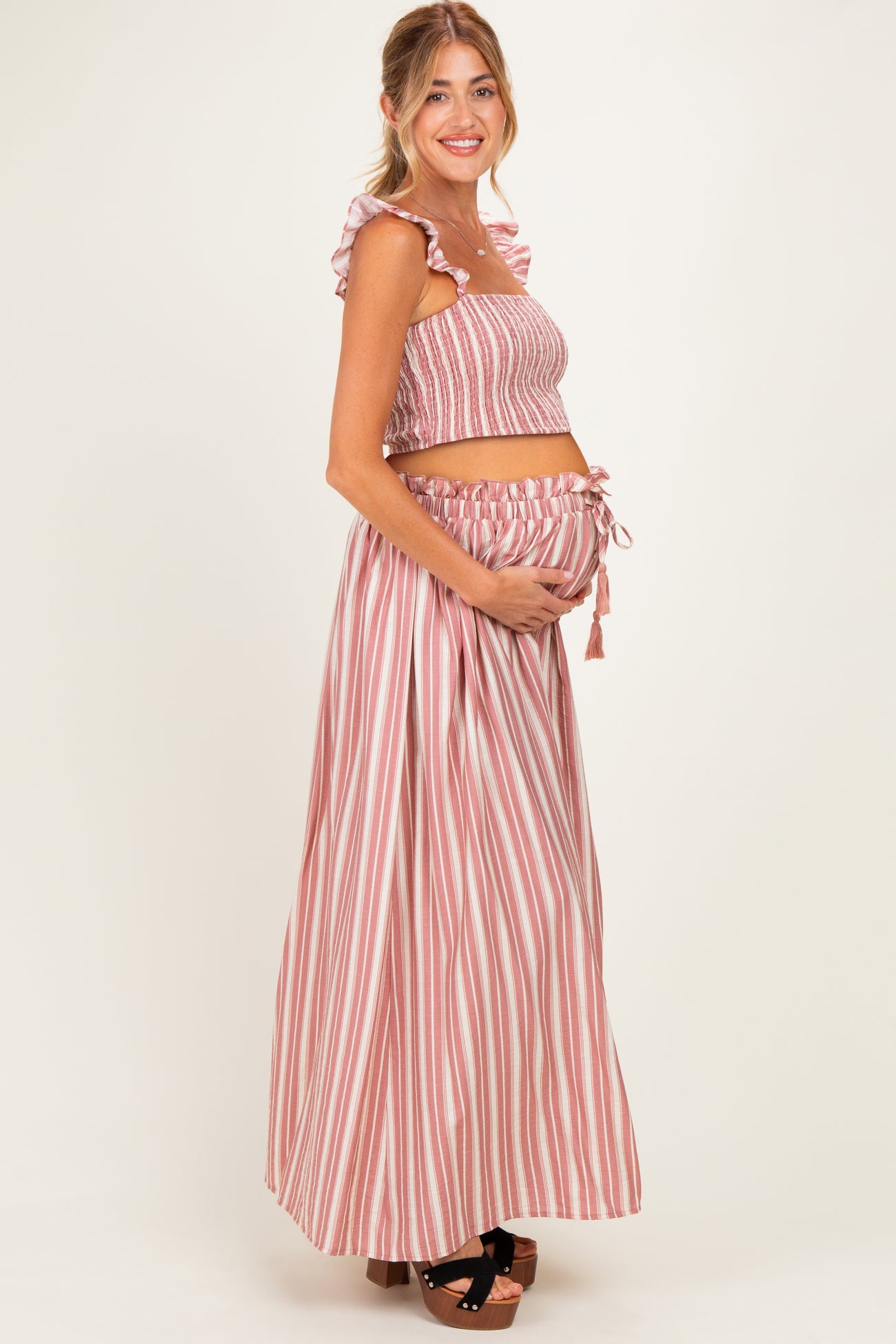 woman wearing a pink and white striped maternity crop top and matching maxi skirt, holding her baby bump