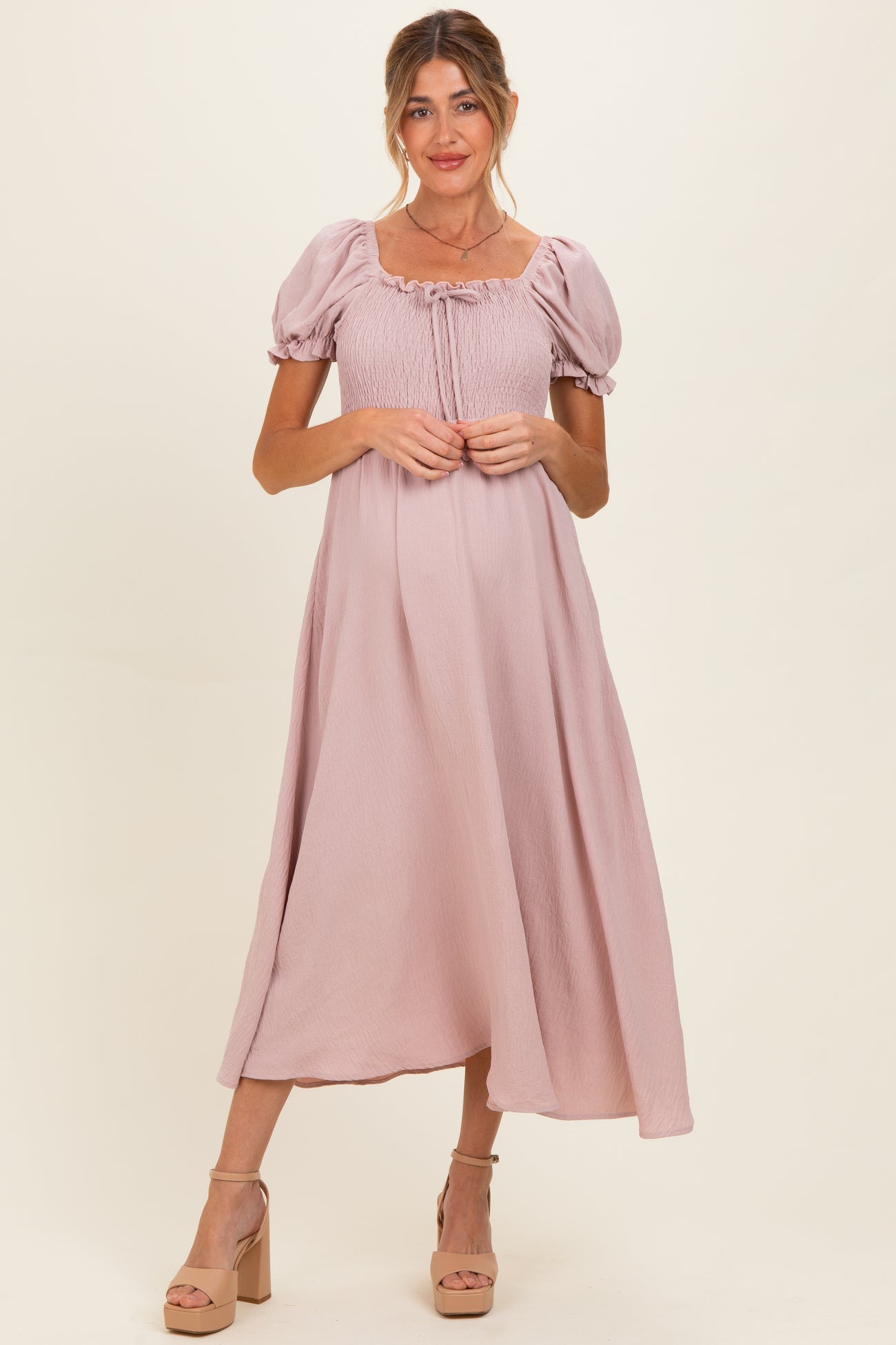 woman wearing a light pink maternity maxi dress with puff sleeves and a smocked bodice, holding her baby bump