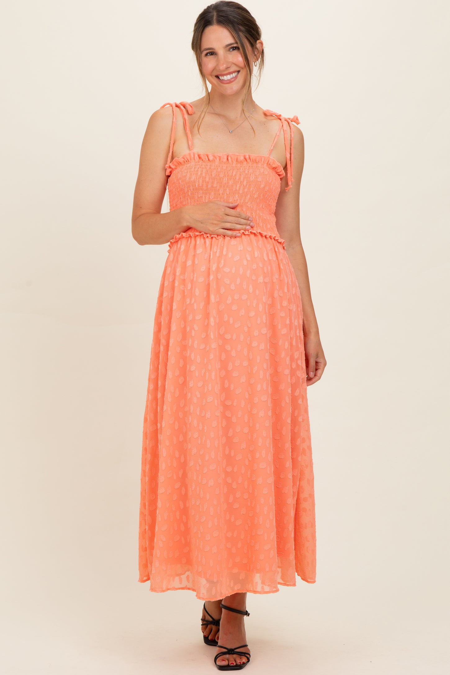 woman wearing a coral maternity maxi dress with adjustable straps, smocked bodice, and holding her baby bump
