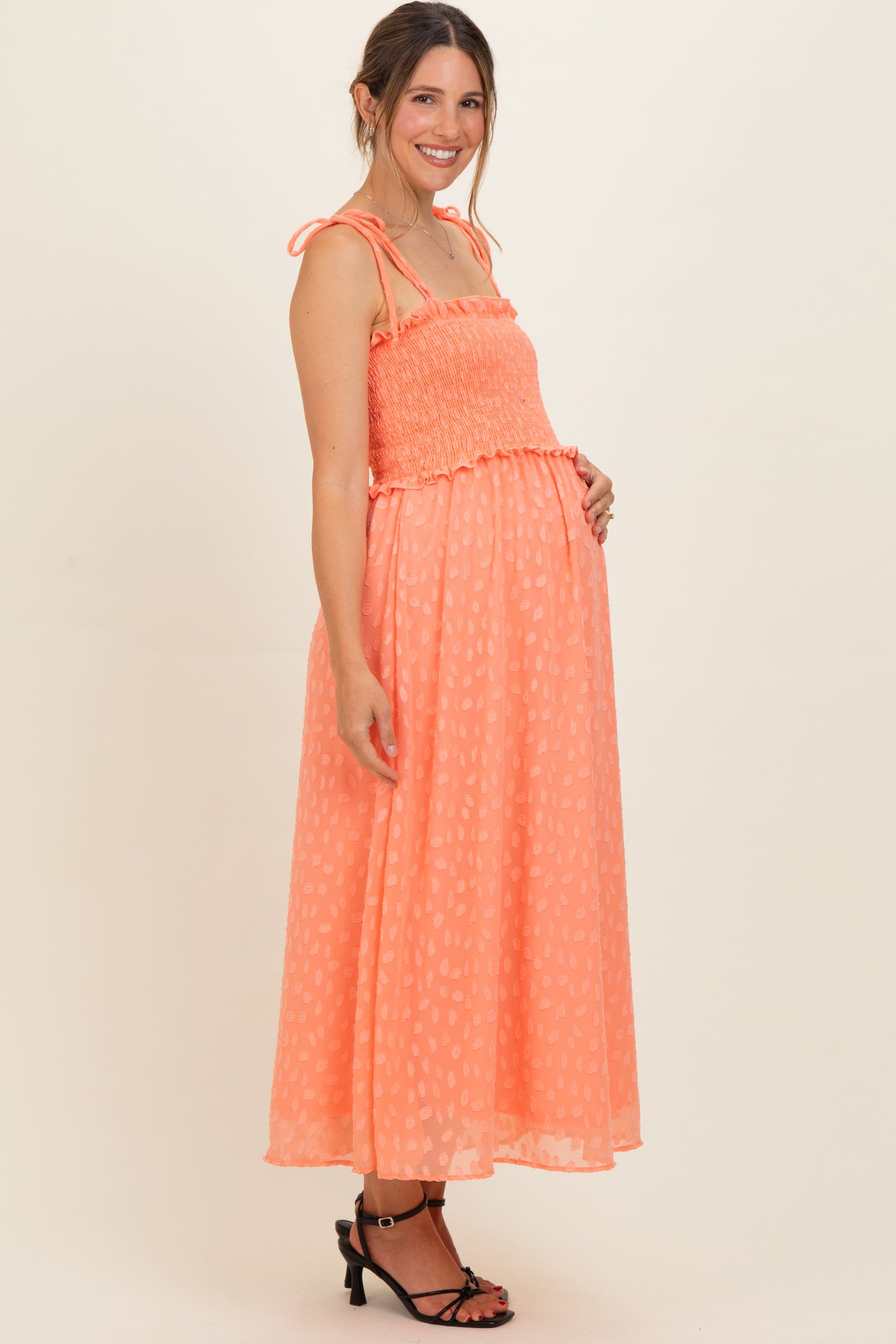 woman wearing a coral maternity maxi dress with smocked bodice and tie straps, showcasing her baby bump