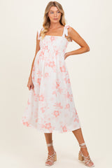 woman wearing a white floral maternity dress with smocked bodice and bow straps, standing with a visible baby bump