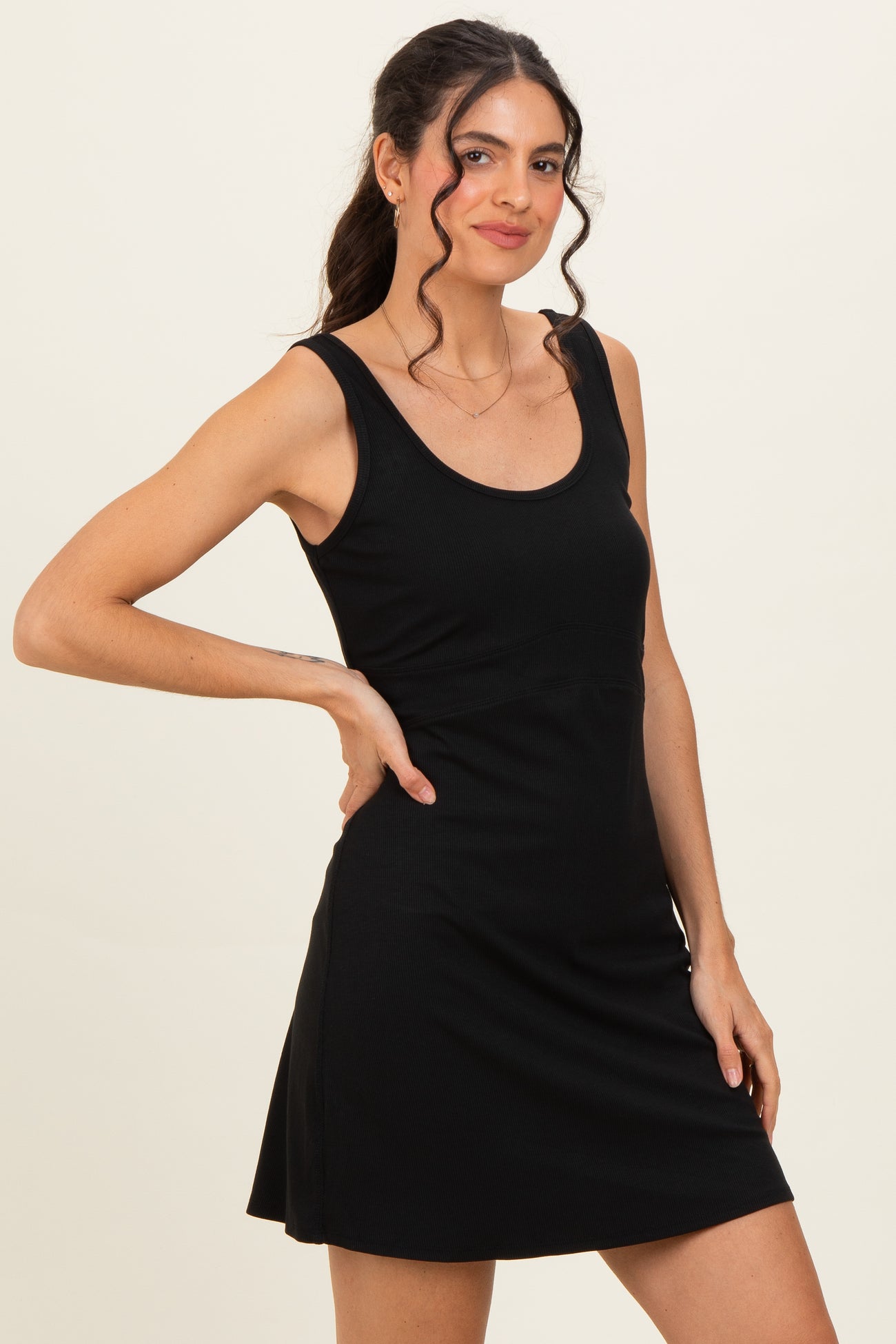 woman wearing a black sleeveless mini dress with a fitted bodice and flared skirt, standing with one hand on her hip