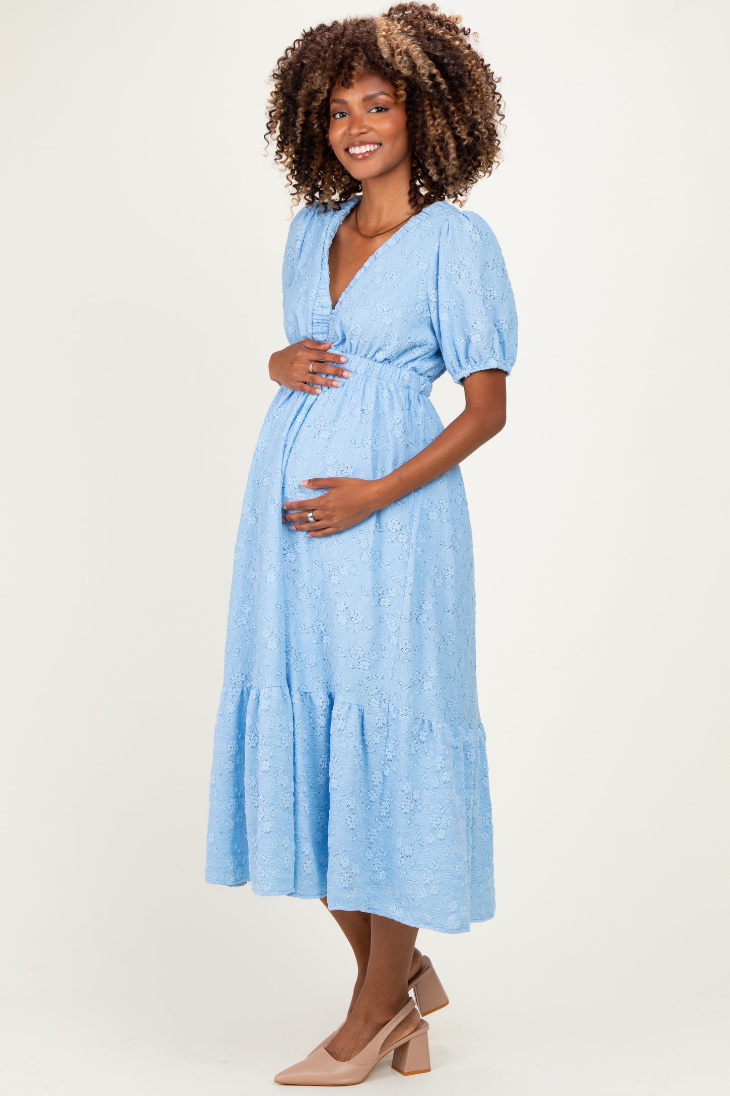 woman wearing a light blue lace maternity dress with puff sleeves and a tiered skirt, holding her baby bump