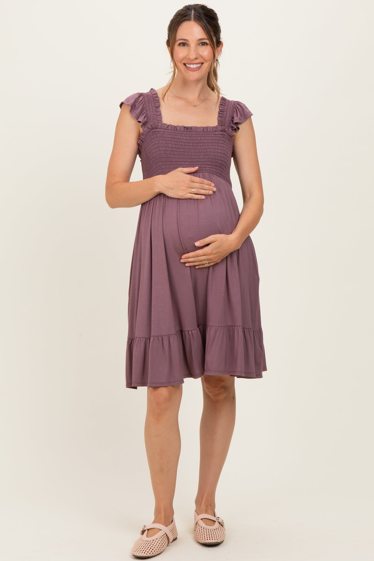woman wearing a mauve maternity dress with ruffled sleeves and a smocked bodice, gently holding her baby bump