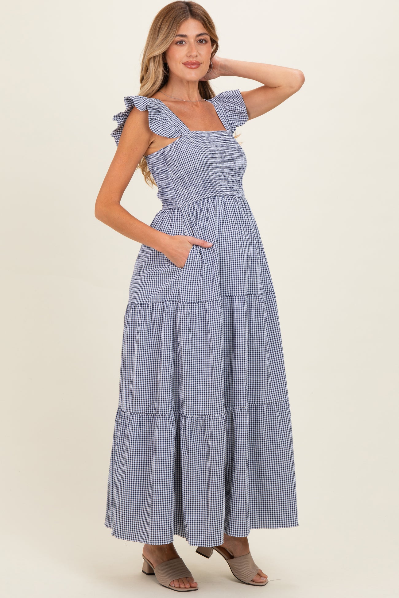 woman wearing a blue and white gingham maternity maxi dress with ruffled straps and pockets, showing her baby bump