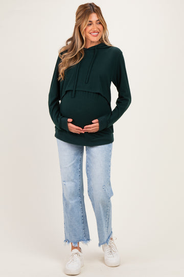 woman wearing a dark green maternity hoodie and distressed light blue jeans, gently cradling her baby bump