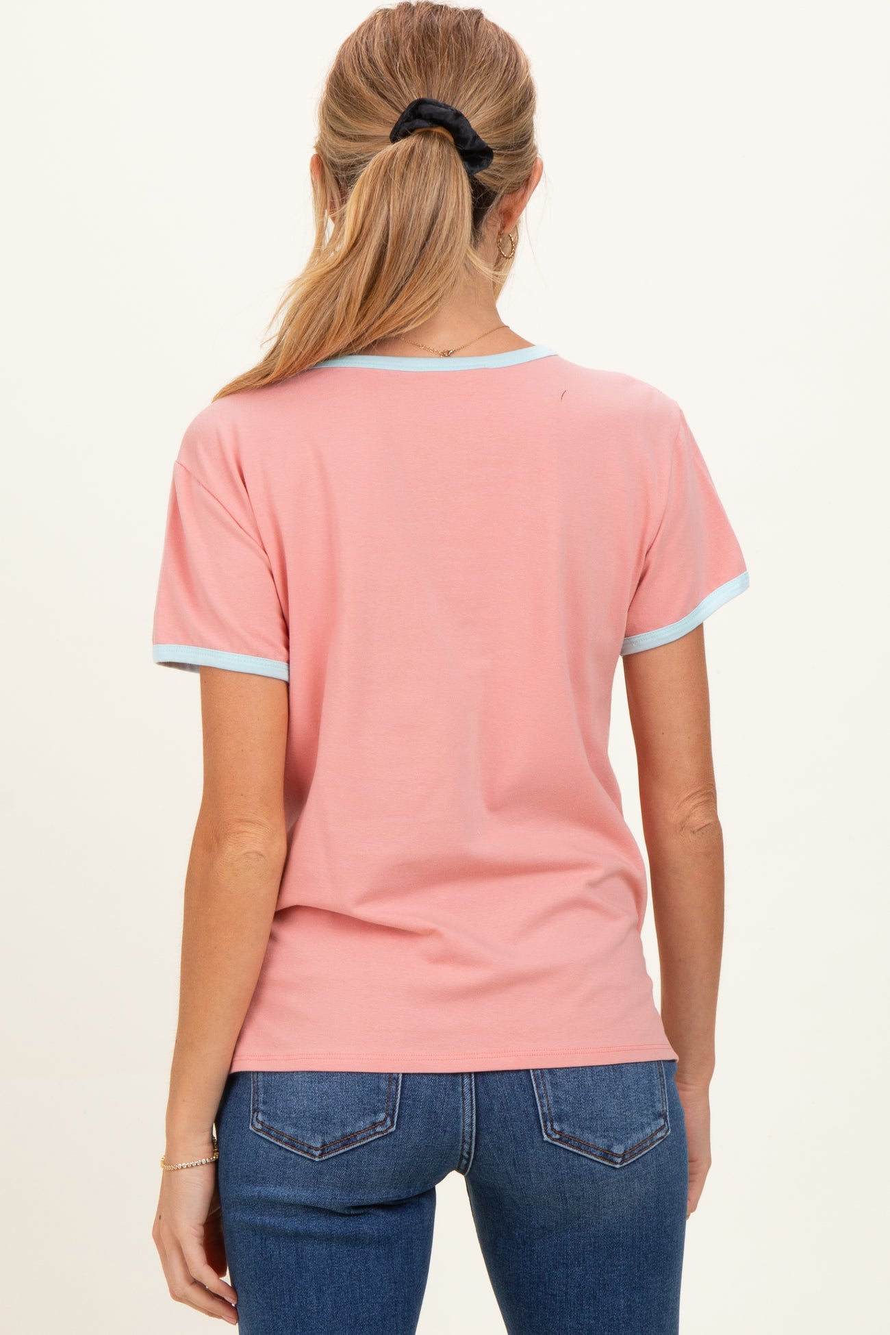 woman wearing a pink maternity t-shirt with light blue trim, viewed from the back while standing against a plain background