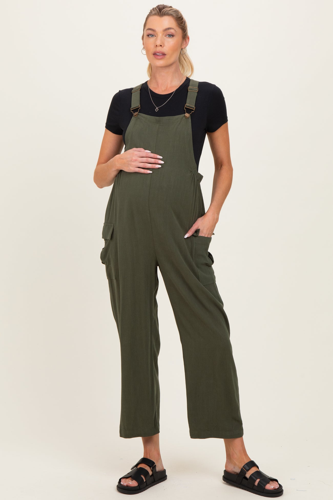 woman wearing a dark green maternity jumpsuit with adjustable straps and pockets, holding her baby bump