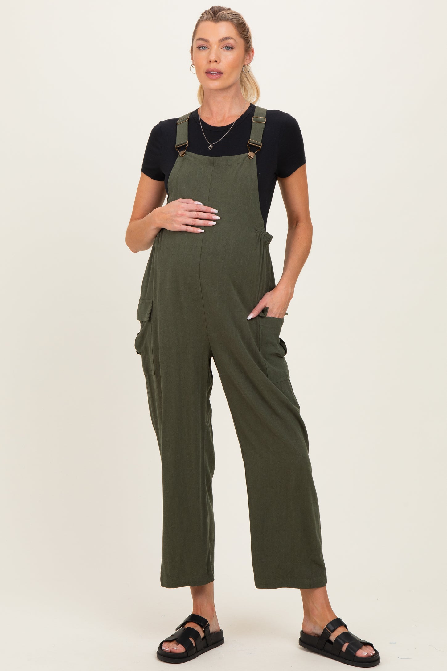 woman wearing a dark green maternity jumpsuit with adjustable straps and pockets, holding her baby bump