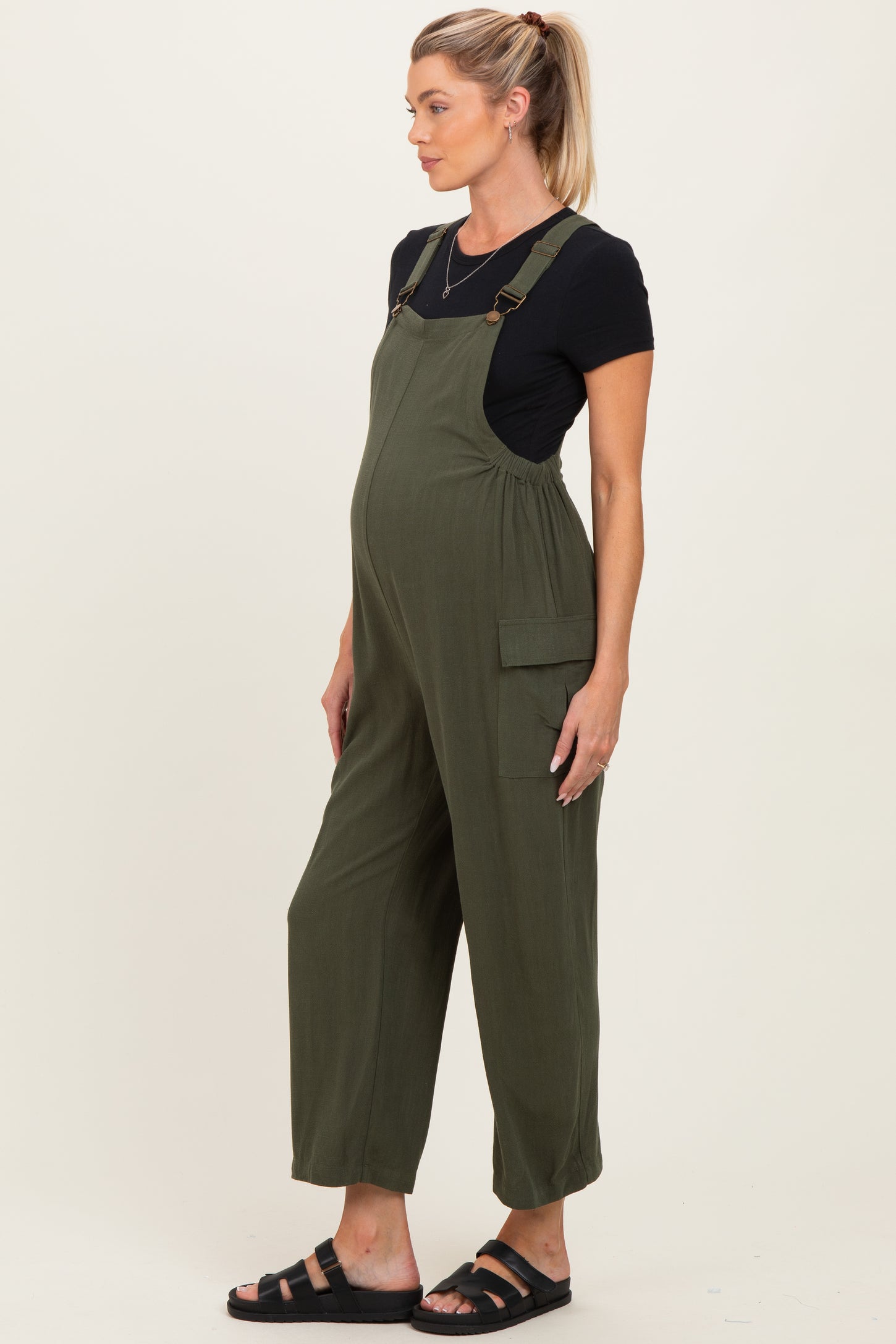 woman wearing an olive green maternity jumpsuit with side pockets and a black t-shirt, showing a baby bump