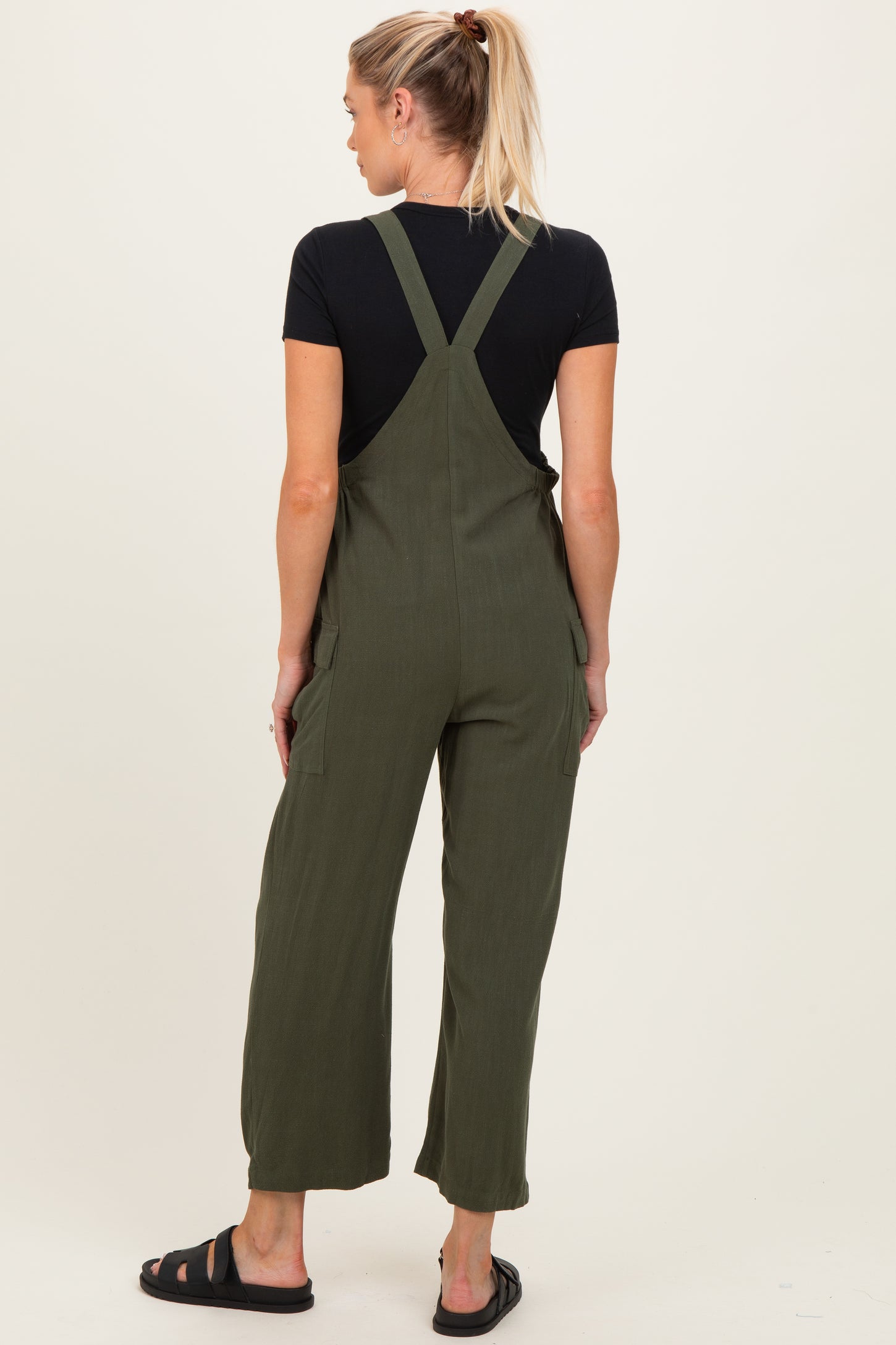 woman wearing olive green maternity overalls with wide legs and adjustable straps, standing with her back to the camera