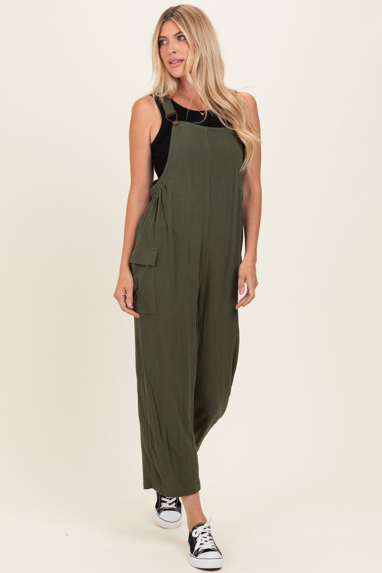 woman wearing an olive green jumpsuit with adjustable straps and side pockets, paired with black sneakers