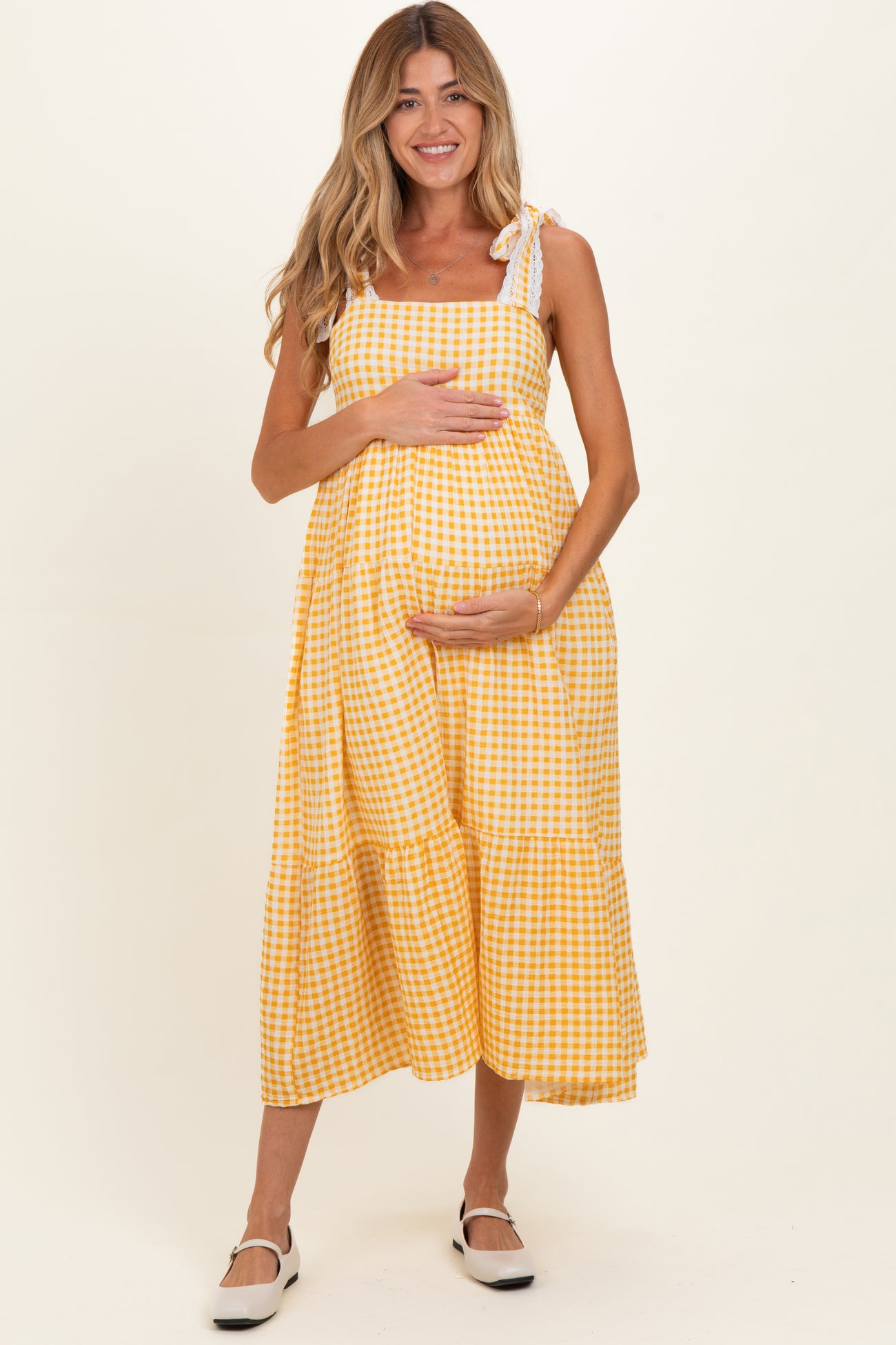 woman wearing a yellow gingham maternity dress with tie straps, cradling her baby bump