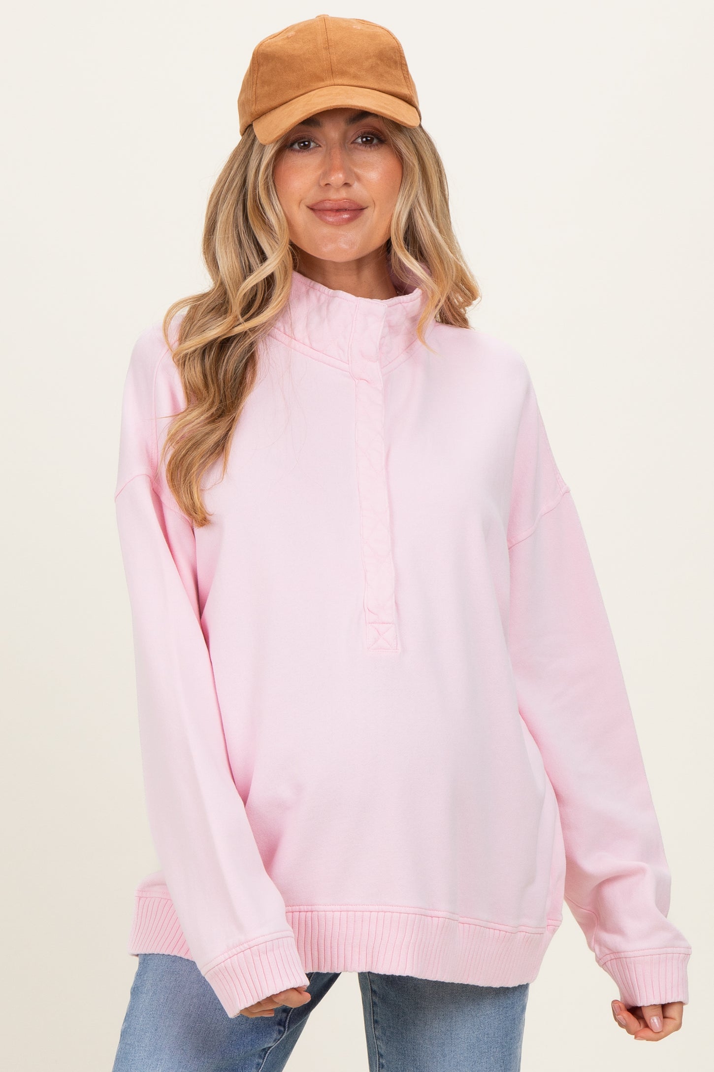 woman wearing a light pink maternity sweatshirt with a high neckline and long sleeves, styled with a tan cap