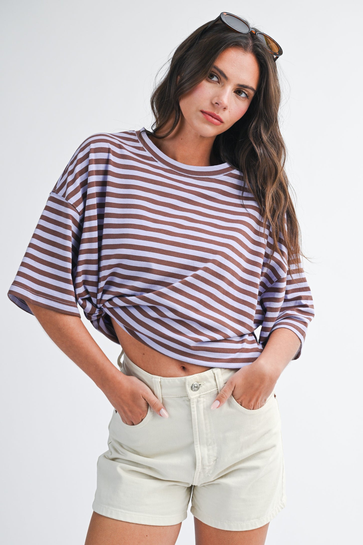 woman wearing a purple and brown striped oversized t-shirt with knotted detail, paired with beige shorts and sunglasses on her head
