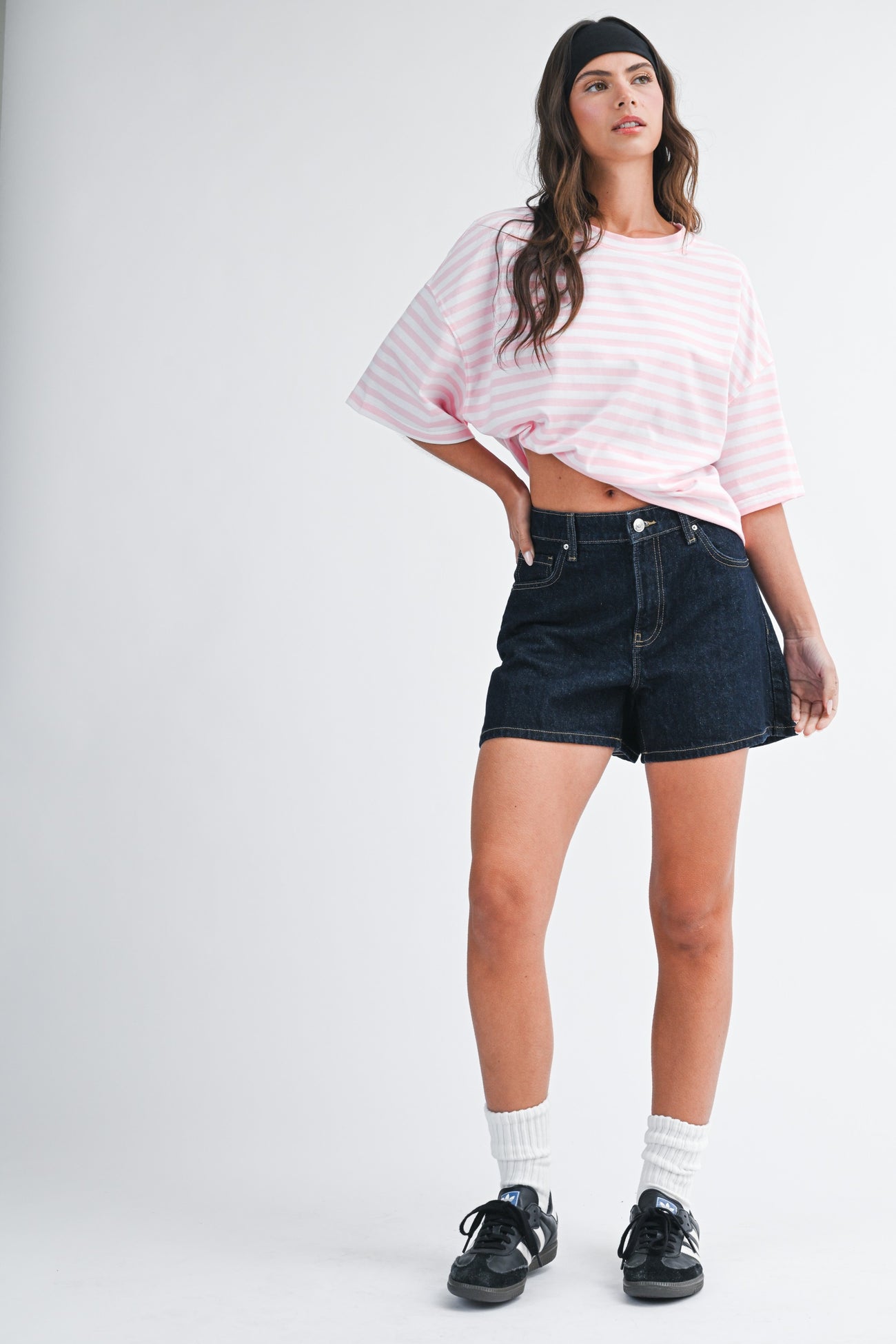 woman wearing a pink and white striped oversized t-shirt and dark denim shorts with a headband and sneakers