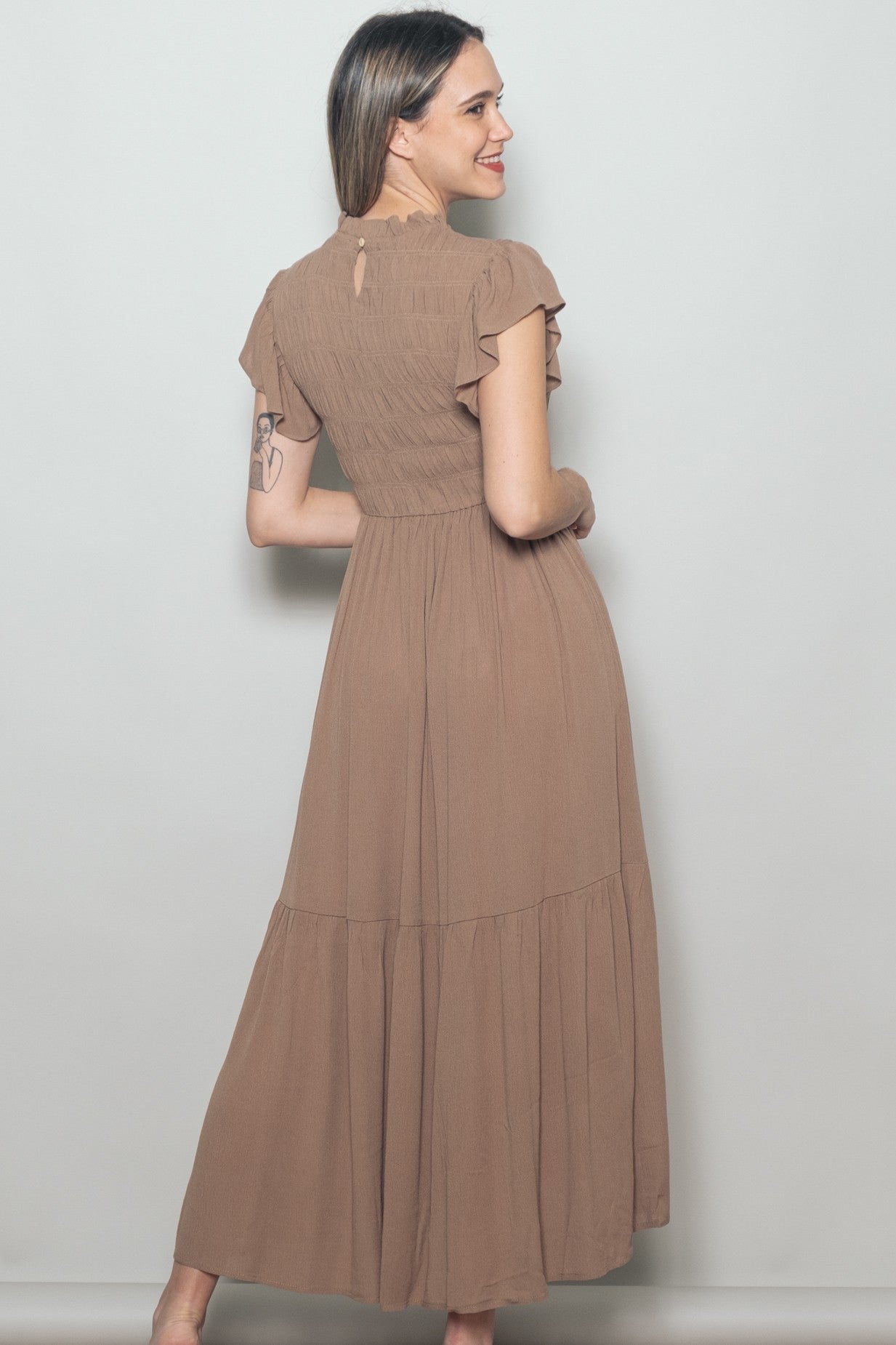woman wearing a brown maxi dress with flutter sleeves and a tiered skirt, standing with her back to the camera