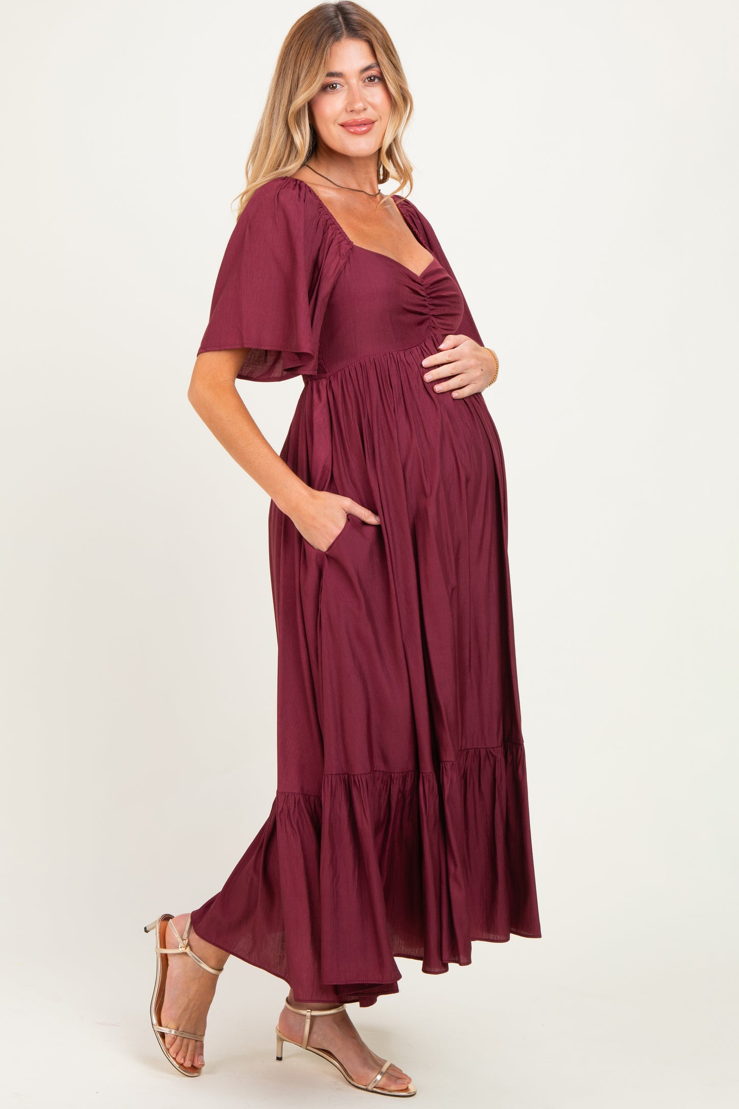 woman wearing a burgundy maxi maternity dress with short sleeves, ruched bodice, and pockets, gently cradling her baby bump