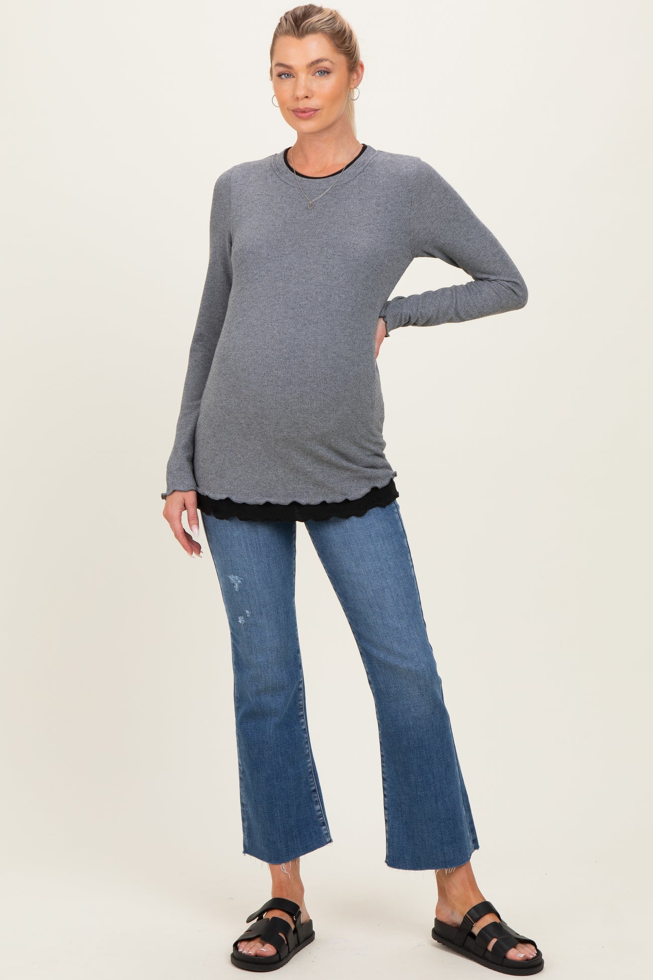 woman wearing a gray maternity top layered over a black scalloped shirt and paired with distressed denim flared jeans and sandals, showing her baby bump