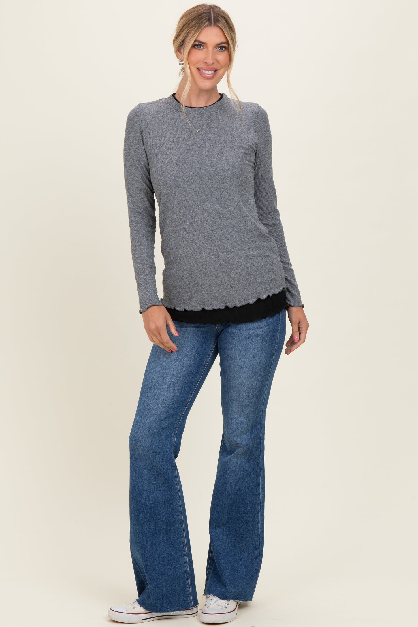 woman wearing a gray long sleeve top with scalloped hem and flared jeans, standing against a light background