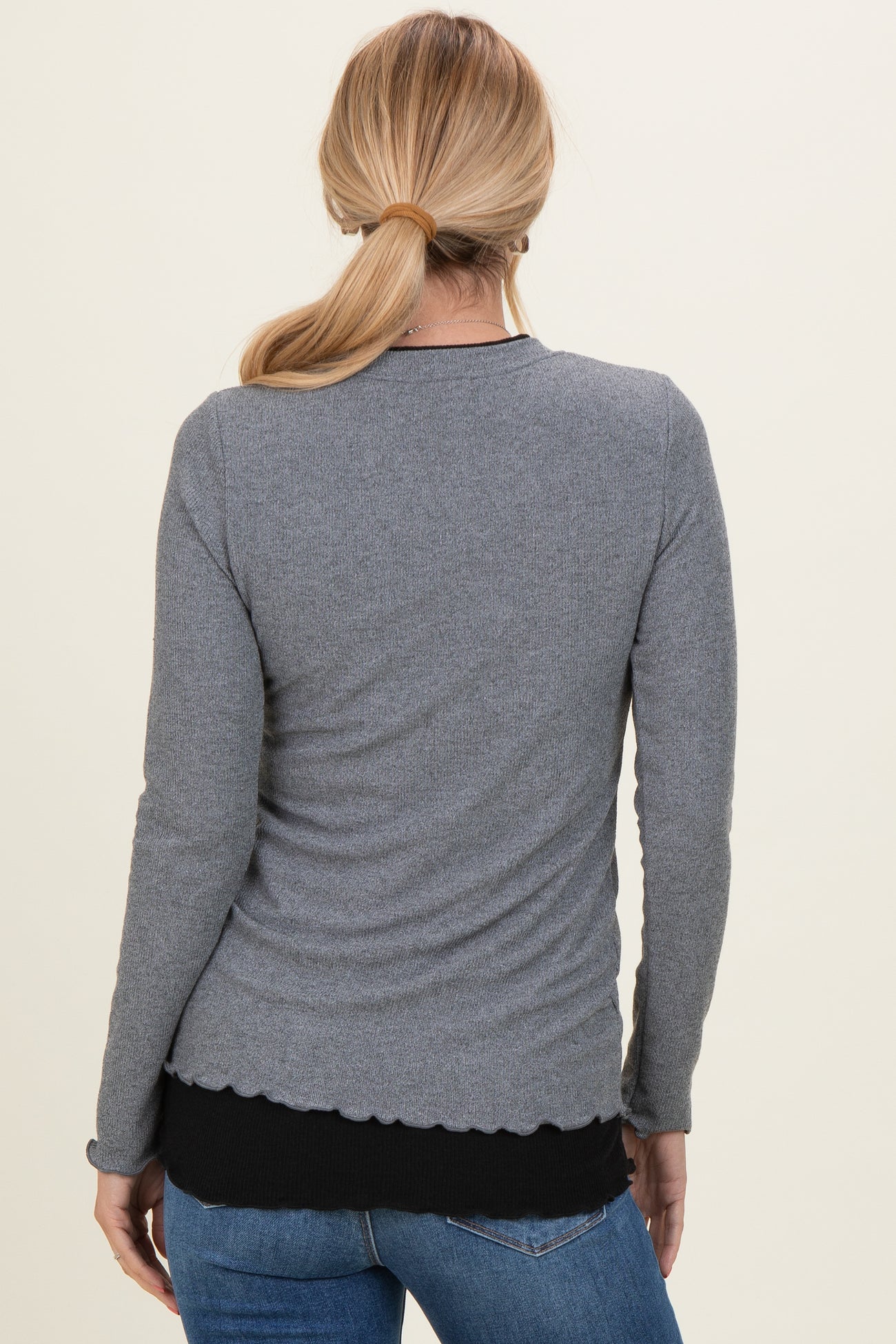 woman wearing a gray long sleeve top with a scalloped hem and styled with dark blue jeans, view from the back