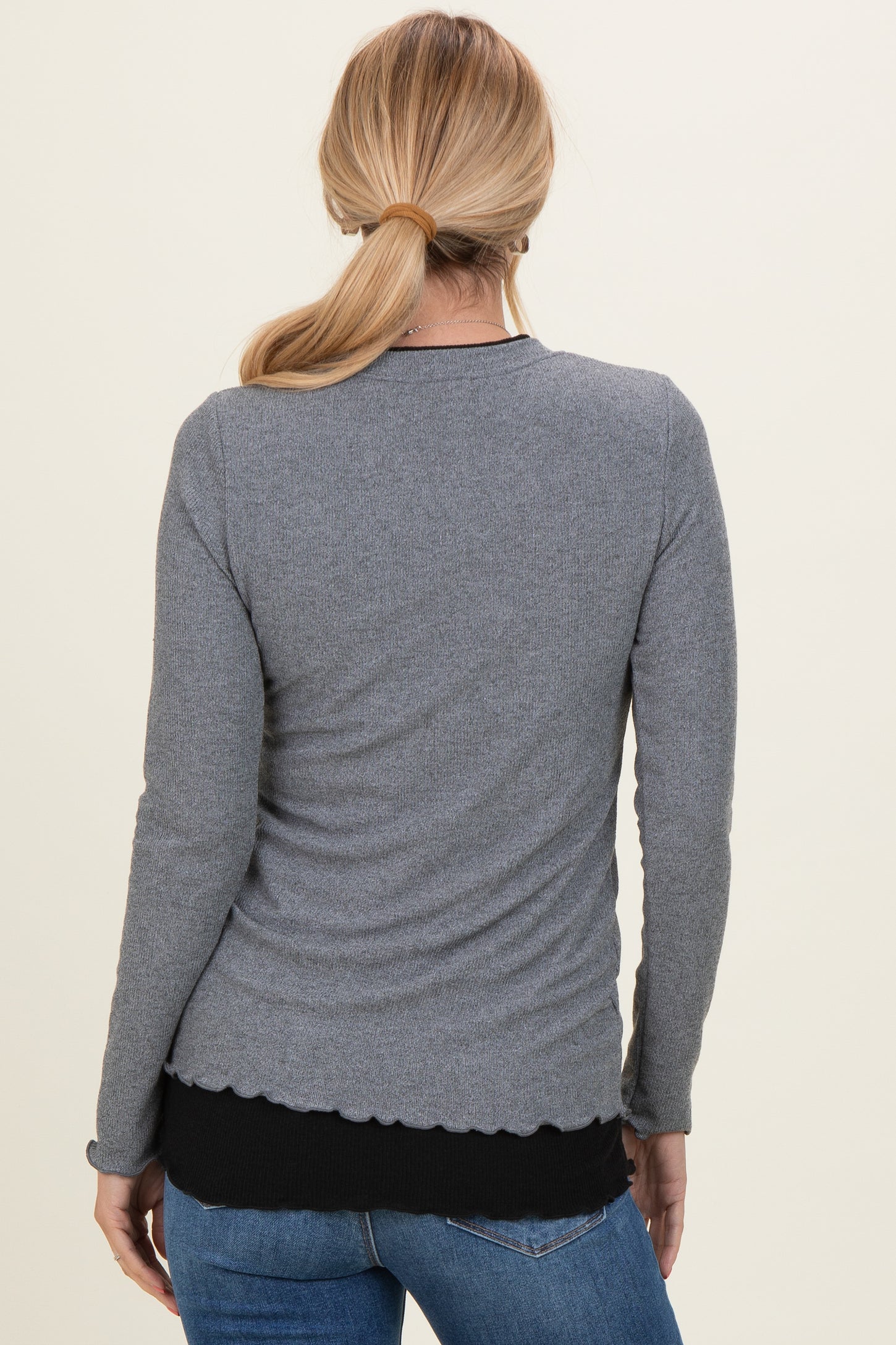 woman wearing a gray long sleeve top with a scalloped hem and styled with dark blue jeans, view from the back