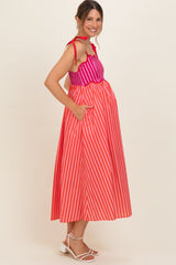 woman wearing a striped maternity maxi dress in red and pink with pockets and adjustable straps, showing her baby bump