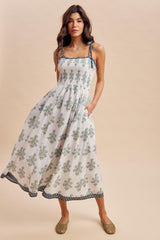 woman wearing a white floral maxi dress with adjustable straps and pockets, standing against a neutral background