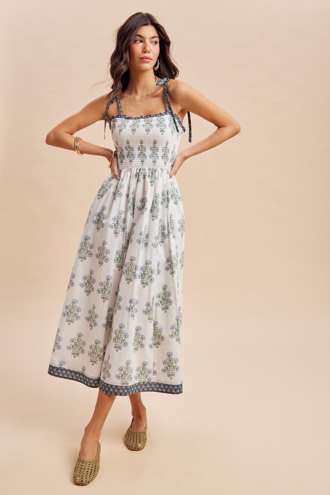 woman wearing a white floral maxi dress with smocked bodice and tie straps, standing with one hand on her hip