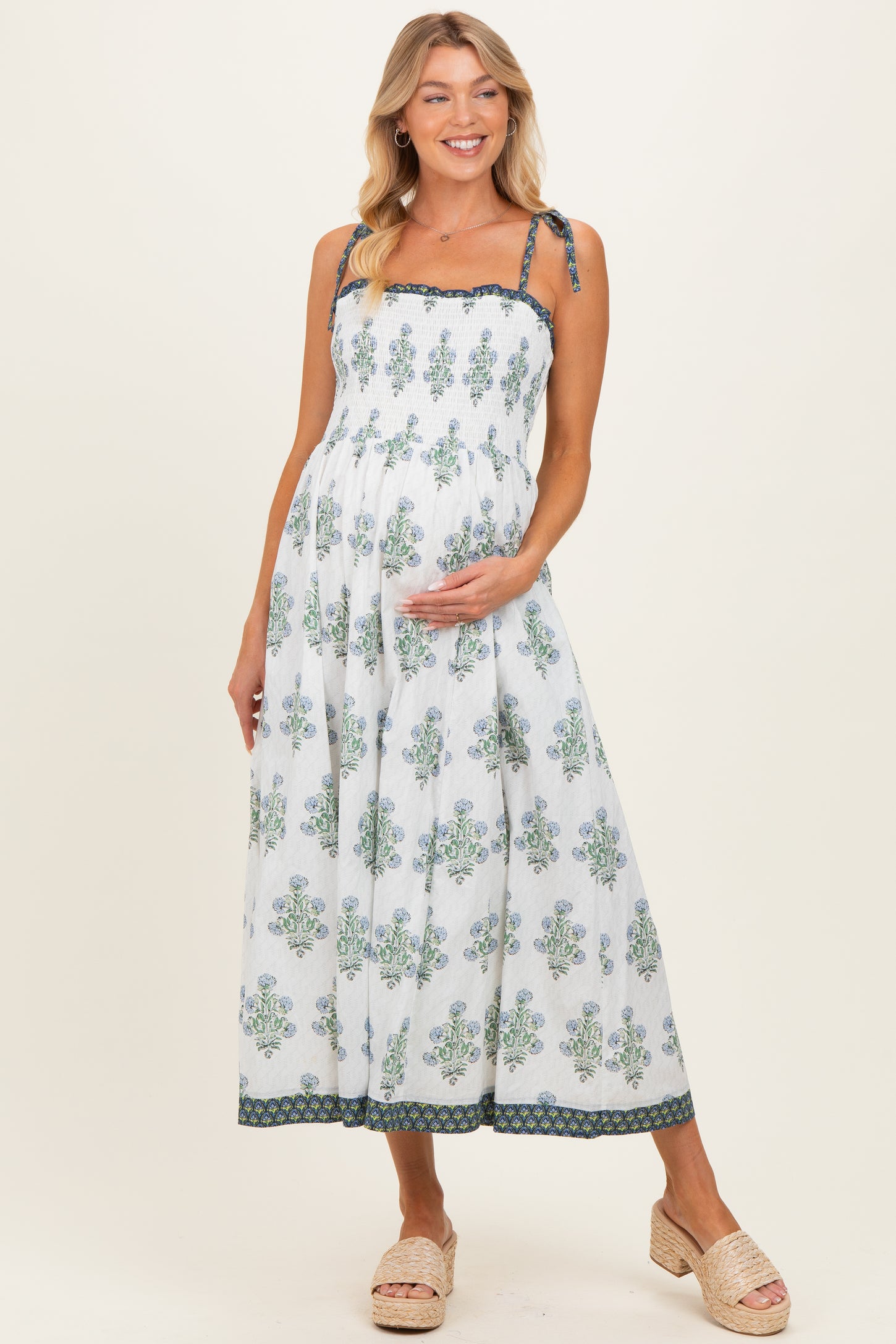 woman wearing a white maternity maxi dress with floral print and tie straps, gently holding her baby bump