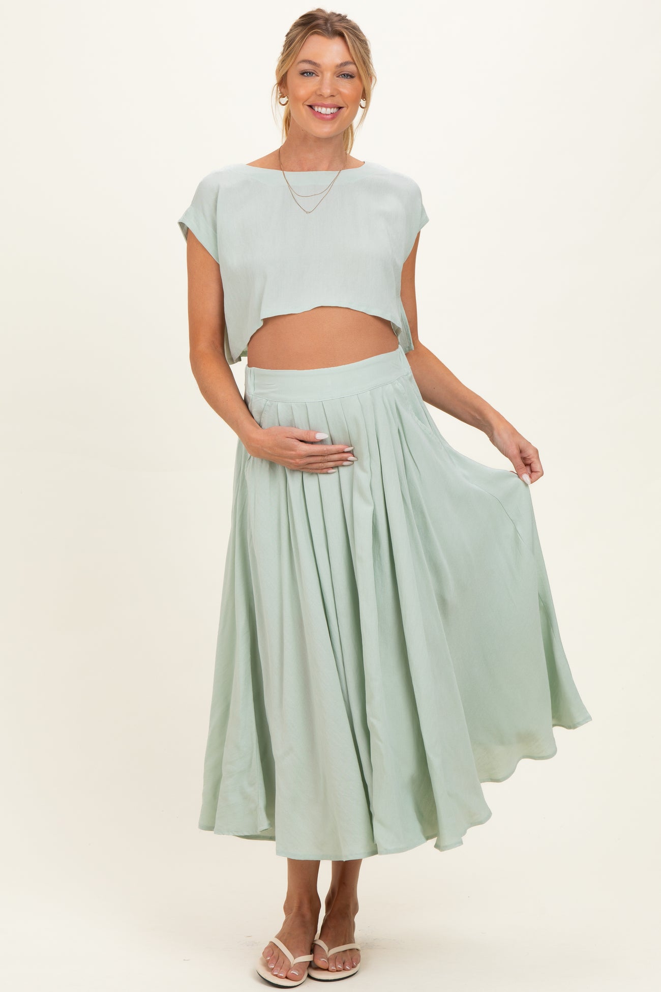 woman wearing a mint green maternity crop top and matching flowy skirt, holding her baby bump