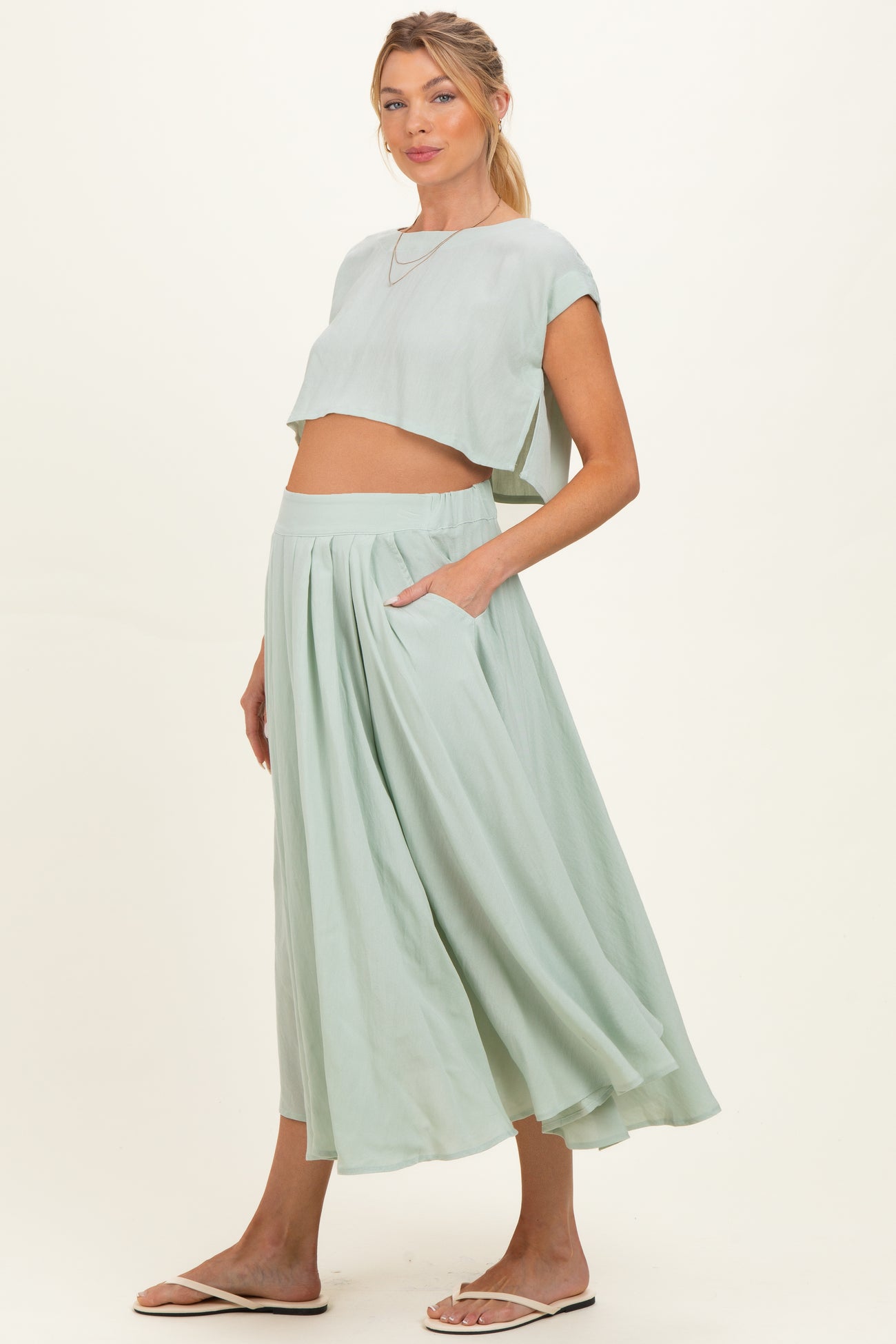 woman wearing a light green maternity crop top and a matching long pleated skirt, showing her baby bump