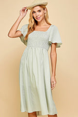 woman wearing a green gingham smocked midi dress with flutter sleeves and a square neckline, holding a straw hat