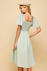 woman wearing a green gingham smocked back dress with short flutter sleeves and a midi length