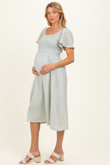 woman wearing a light green maternity dress with short puff sleeves and smocked bodice, holding her baby bump