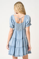 woman wearing a light blue tiered denim dress with short puff sleeves and a tie-back detail