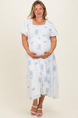 woman wearing a white maternity plus dress with blue floral patterns and holding her baby bump