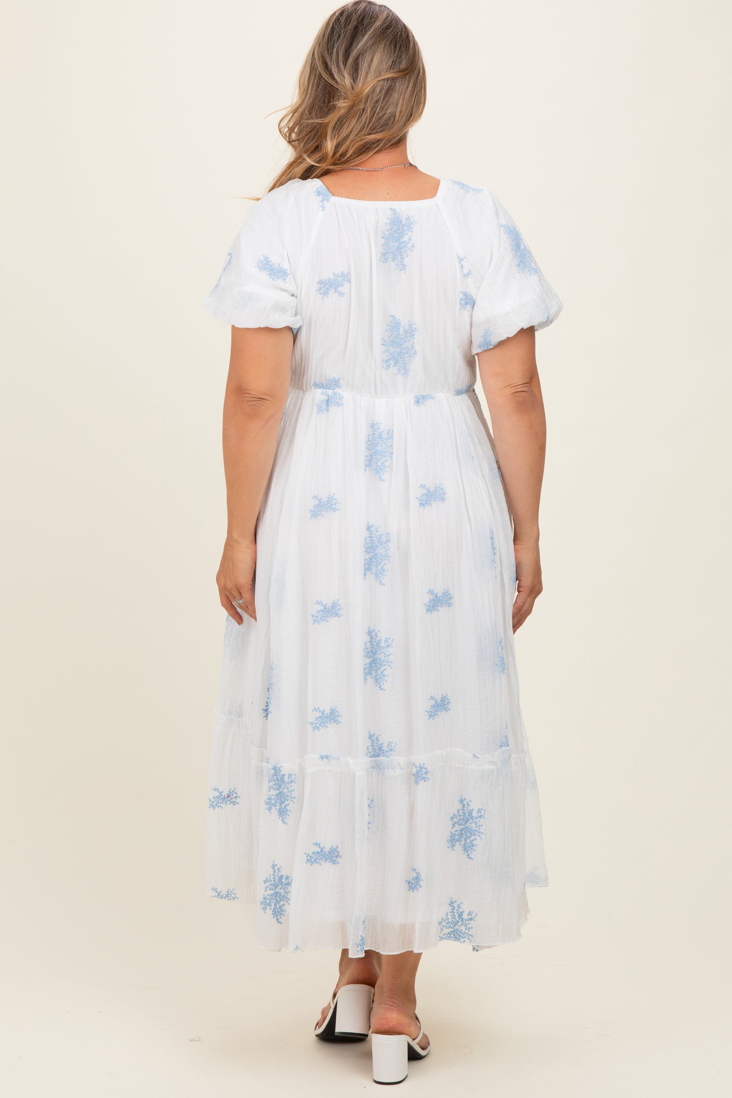 woman wearing a white maternity plus dress with blue floral embroidery and puff sleeves, showing a baby bump from the back