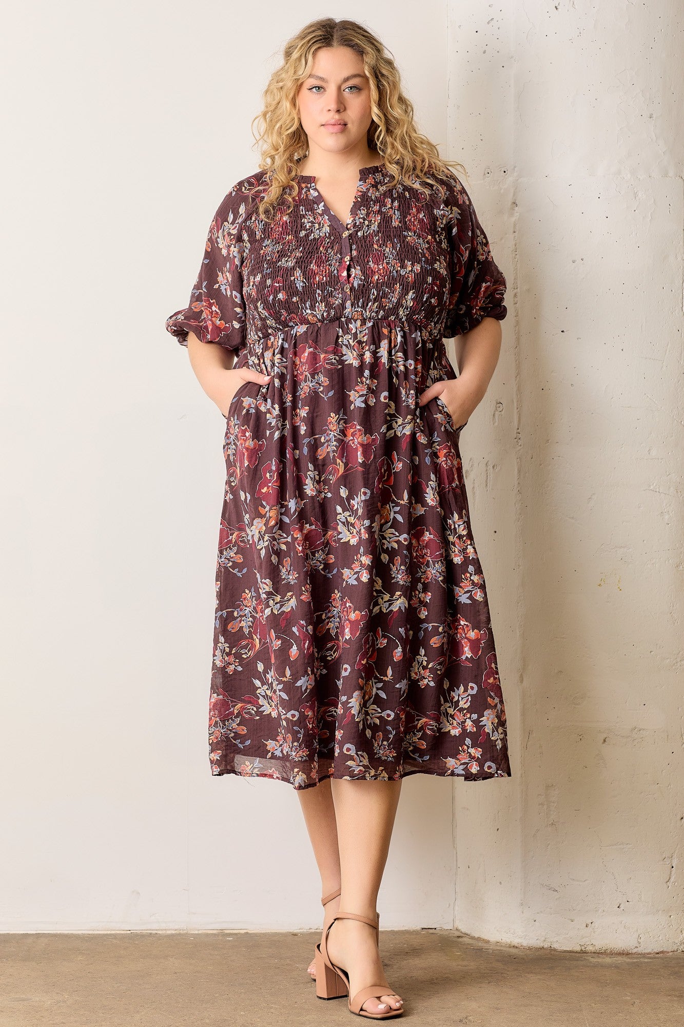 woman wearing a burgundy floral midi dress with smocked bodice and pockets, standing against a white wall