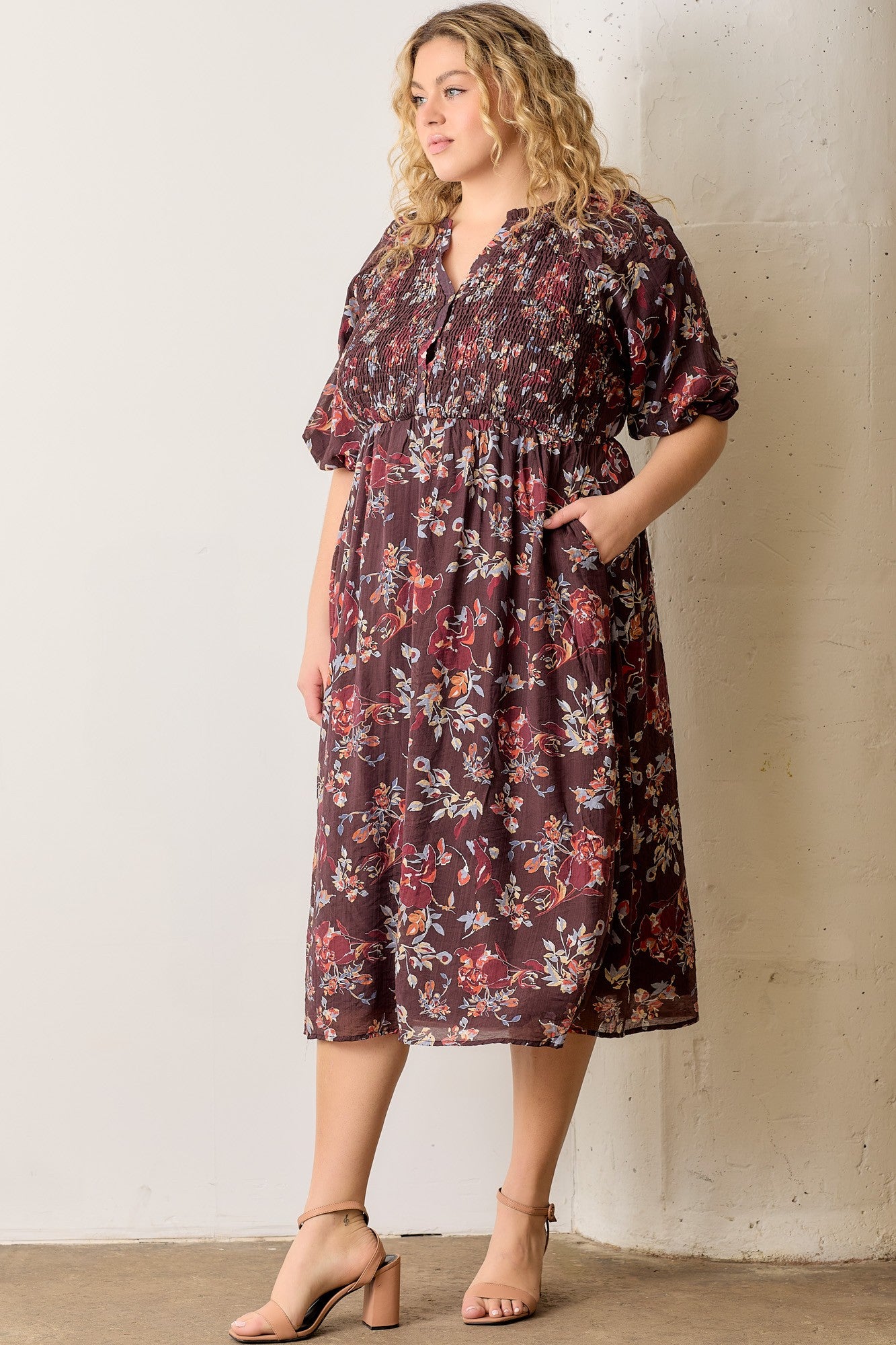 woman wearing a burgundy floral plus size dress with smocked bodice and short sleeves, standing against a wall