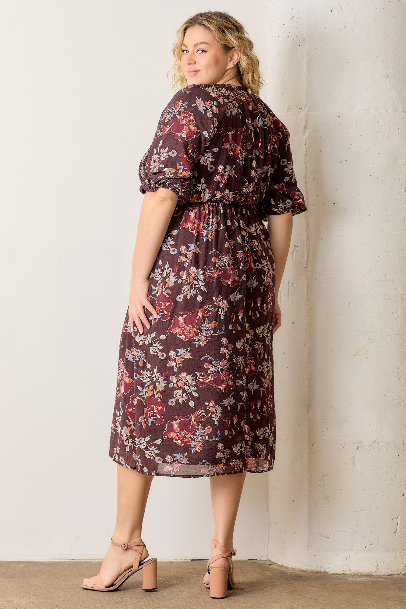 woman wearing a burgundy floral plus-size midi dress with puff sleeves and a cinched waist, standing sideways against a wall