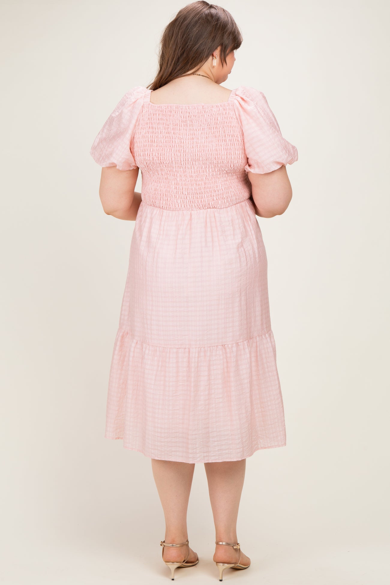 woman wearing a pink checkered smocked maternity dress with puff sleeves and a tiered hem, viewed from the back