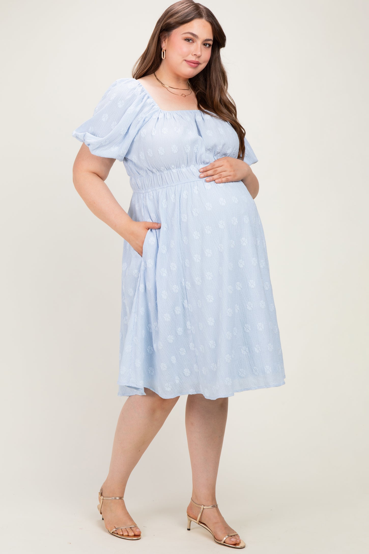 woman wearing a light blue floral maternity dress with short puff sleeves and pockets, holding her baby bump