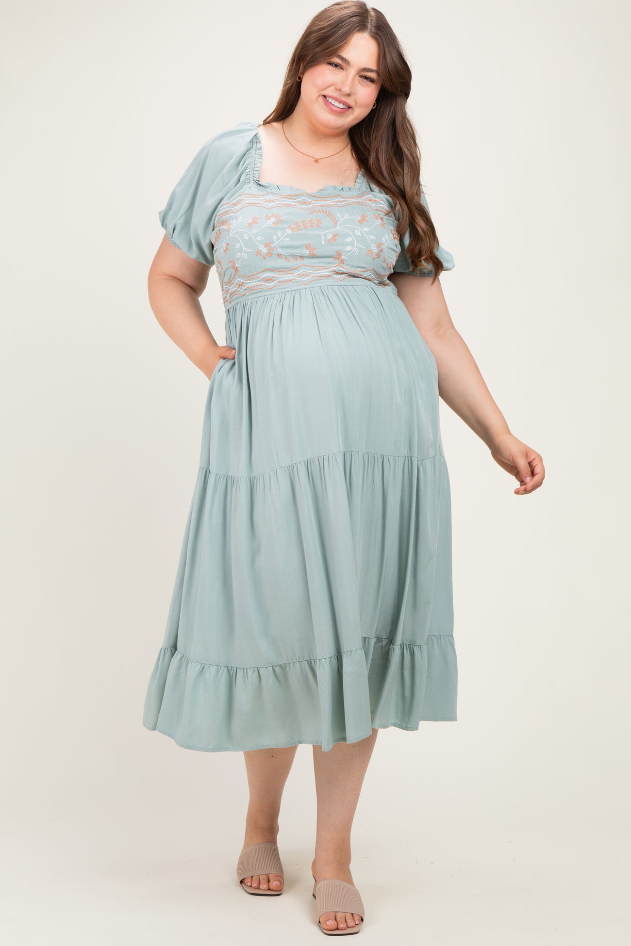 woman wearing a sage green maternity plus maxi dress with embroidered bodice and short puff sleeves, holding pockets