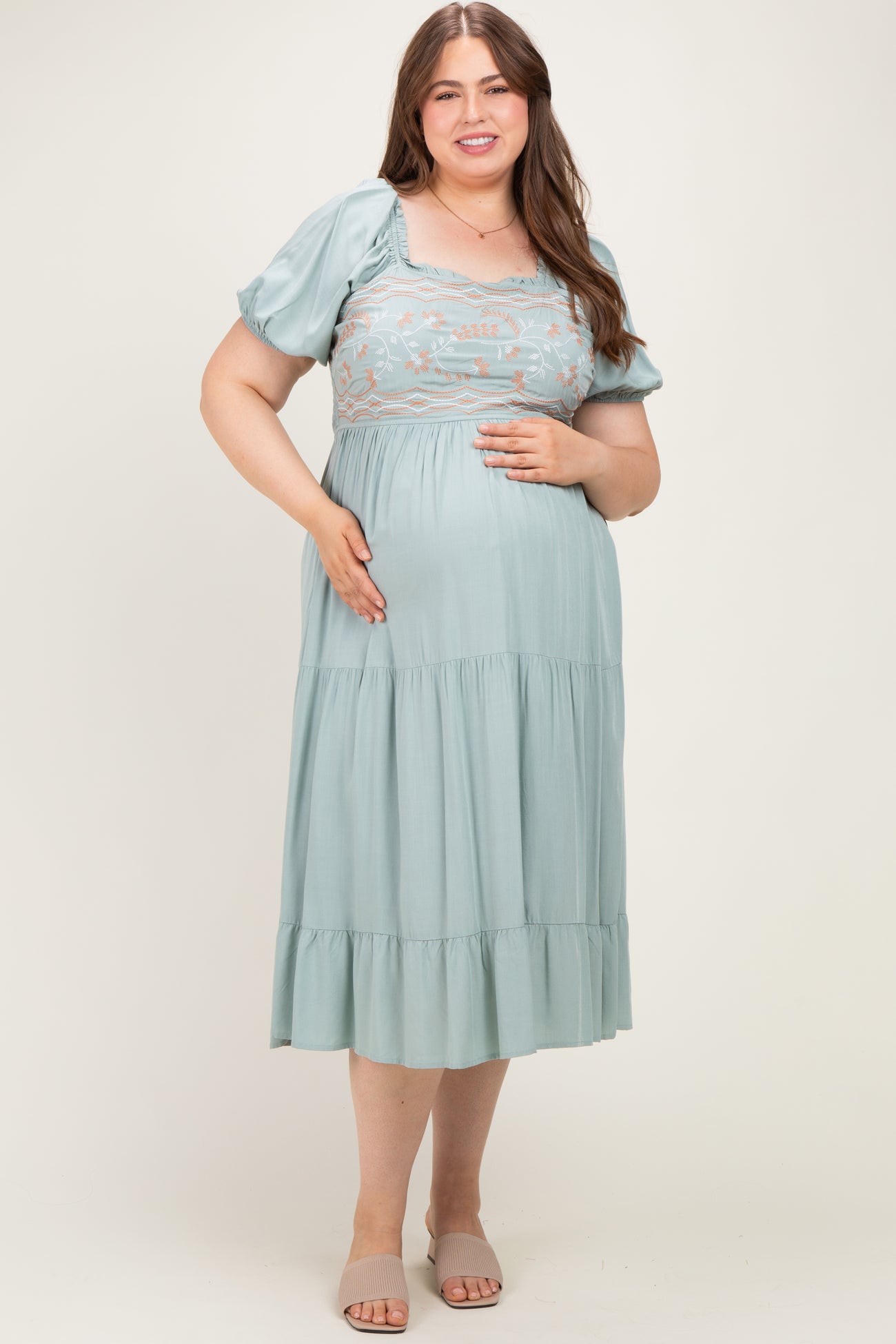woman wearing a light green maternity plus dress with puff sleeves and embroidered bodice, holding her baby bump