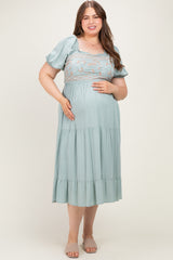 woman wearing a light green maternity plus dress with puff sleeves and embroidered bodice, holding her baby bump